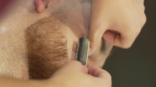 The barber carefully shaves the client's beard using a special shaving razor. It is a close up video