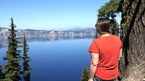 Mujer de pie junto a un árbol con vistas al lago del cráter con el pico de la montaña en la distancia