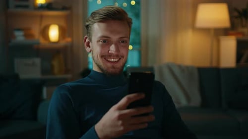 Handsome Man Relaxes at Home Uses Smartphone. In the Background Cozy Living Room in the Evening wit