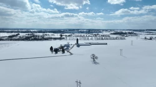 Rural farmland covered in snow on sunny winter day. Vast landscape. Aerial flight over large farm.
