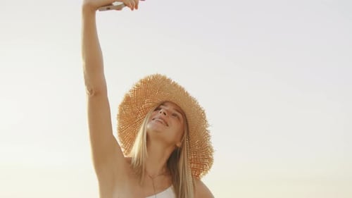 Young Woman Enjoys Golden Hour at Beach While Taking a Selfie Wearing a Stylish Straw Hat Capturing