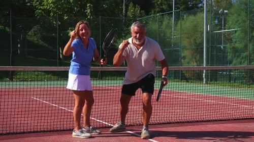 senior couple celebrates tennis victory, shouting and raising their fists while holding racquets