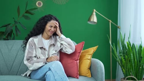 Una joven infeliz sentada en el sofá se frota la sien con los ojos cerrados y sufre un dolor de cabeza doloroso