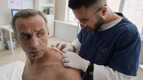 Young Male Doctor Examining Skin Rash of Middle-Aged Man in Clinic