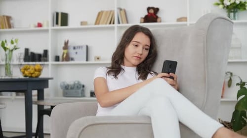 Pretty young girl sitting in the chair indoors and using smartphone