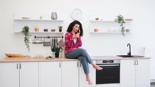 Lächelnde Frau sitzt auf einer Küchentheke, hält Kaffee in der Hand und telefoniert