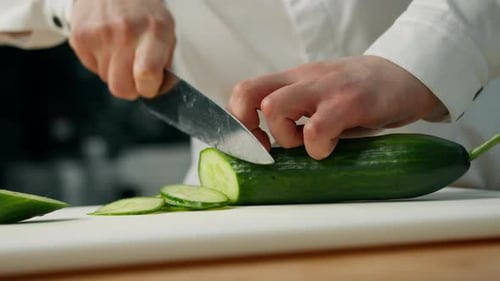 professional kitchen: chef shallowly cuts a cucumber to prepare a burger or salad
