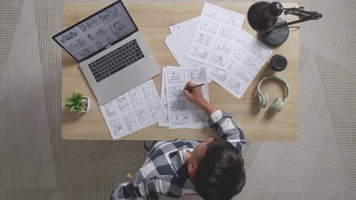 Ansicht von oben auf einen asiatischen männlichen Künstler, der Rückenschmerzen hat, während er mit einem Laptop ein Storyboard auf dem Tisch zeichnet