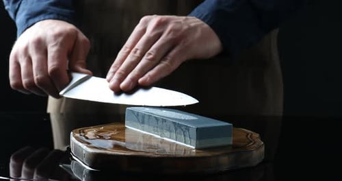 Man sharpening knife with sharpener at mirror table against black background, closeup