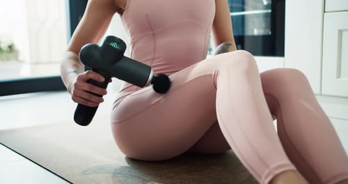 Unrecognizable Woman in Sportswear Sitting on Mat and Massaging Leg with Massage Percussion Device