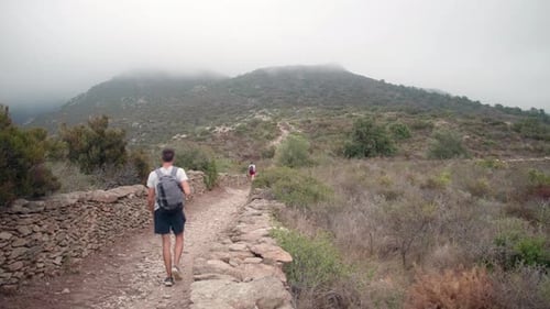 Couple Of Hikers With Backpack Walking At Dirt Path Along Countryside Fields With Misty Mountain In