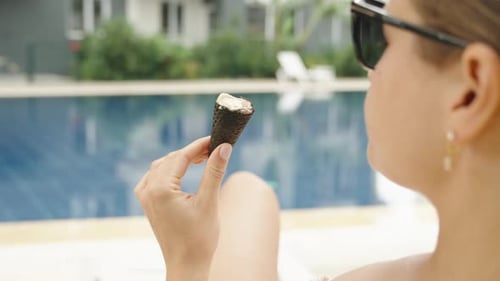 A young woman in a swimsuit is lying by the pool and eating a black ice cream cone.
