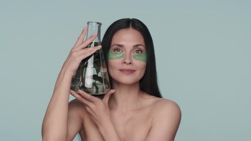 Uma mulher com manchas verdes de hidrogel sob os olhos segura um frasco com uma planta seminua no