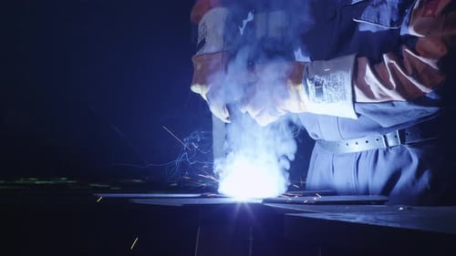 Heavy industry worker at a factory is welding metal in dark interior.