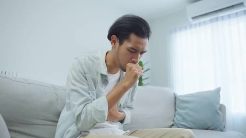 Hombre guapo asiático que se siente mal y enfermo, tosiendo en la sala de estar.