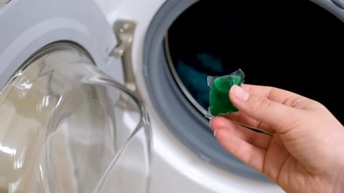 Female hand holding capsule with green liquid powder against background of washing machine close-up.
