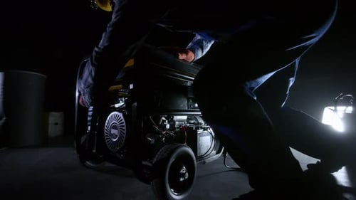 A Man in Blue Overalls and a Yellow Helmet in the Dark Turns on a Gasoline Generator and Lights Up