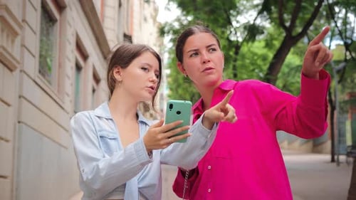 Dos mujeres jóvenes que señalan hacia adelante disfrutando de un momento juntas al aire libre, sonriendo y comprometiéndose en una ciudad
