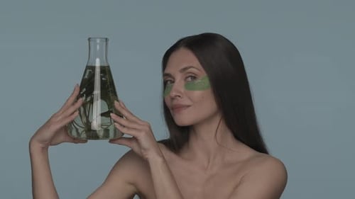 Uma mulher com manchas verdes de hidrogel sob os olhos segura um frasco com uma planta seminua no