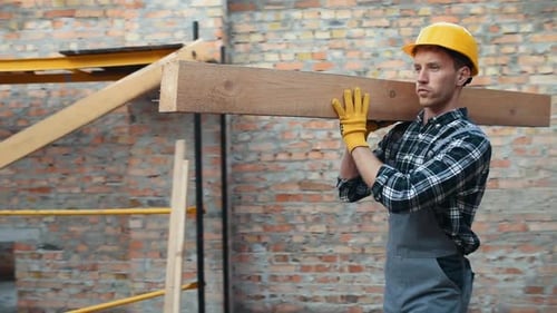 Walks with piece of board. Construction worker in uniform and safety equipment have job on building
