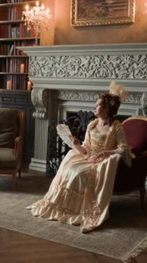 Aristocratic Woman in Ornate Silk Dress Sitting by Fireplace and Waving Fan