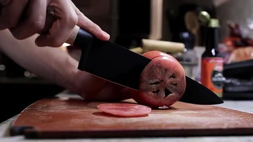 Caucasian male hand slices red raw tomato on chopping block with sharp knife, static side close up