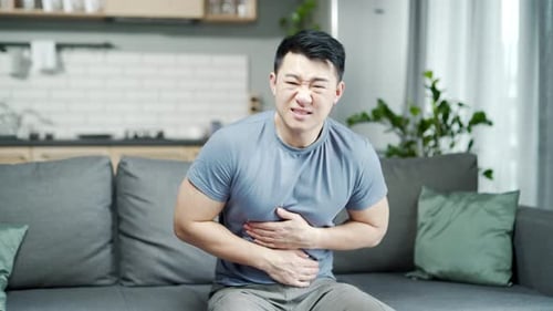 Asian man suffers from abdominal pain while sitting at home on the couch. stomach ache. diarrhea,