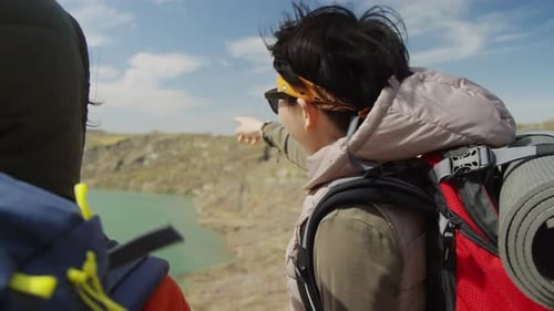 Dos turistas de pie en la ladera rocosa apuntando al lago, viento fuerte
