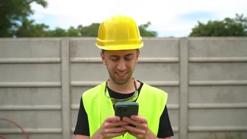 General Labourer In Hard Hat And Safety Vest Typing On Smartphone Outdoor. medium shot