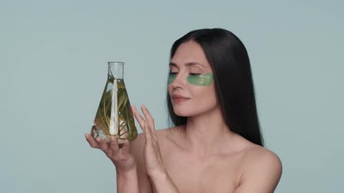 Uma mulher com manchas verdes de hidrogel sob os olhos segura um frasco com uma planta mulher seminua no