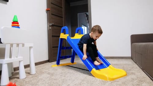 Active toddler boy playing on the slide in his room. Cute baby climbs up backwards.
