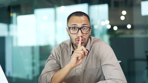 Young man holds index finger near lips while sitting at workplace at desk in modern office. Handsome