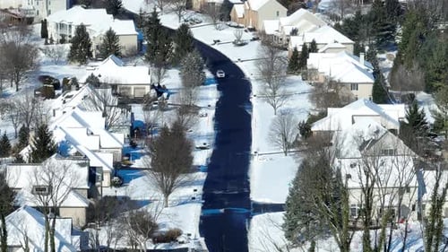 Aerial shot of a singular vehicle driving down a neighborhood street. Winter scene with snow on hous