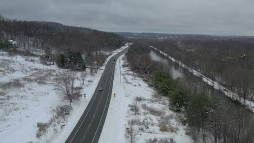 Flying over trees as multiple vehicles travel along highway next to river in winter on overcast day.
