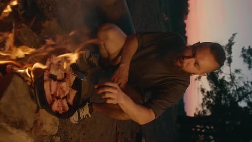 Attractive Young Man with a Beard on a Picnic By the River Fries Meat in a Pan on a Fire