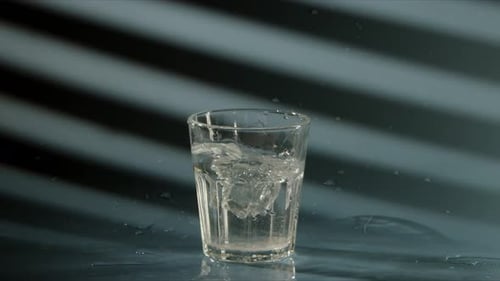 Piece of Crushed Ice Falls Into Transparent Glass with Clean Drinking Water on Blue Background