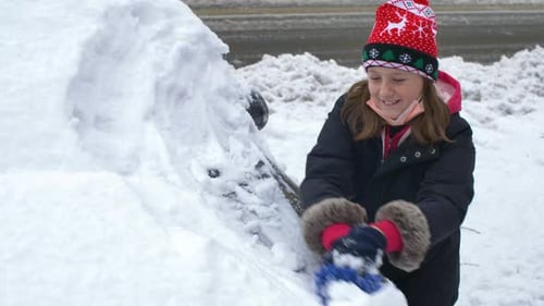 young girl helps clear snow from a car in winter after a snowstorm in a parking lot.