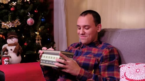 Sad Man Lying on a Sofa Near the Christmas Tree, Opening Gift Disappointed and Unhappy