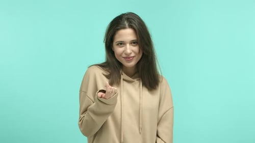 Hermosa mujer caucásica en sudadera con capucha beige coqueteando y sonriendo, moviendo el dedo mostrando más cerca