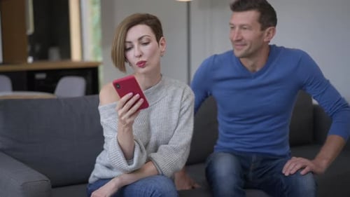 Beautiful Woman Texting on Smartphone Ignoring Man Sitting Down on Couch with Shocked Facial