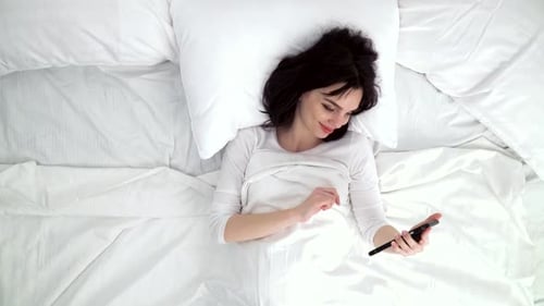Woman With Mobile Phone Lying In Bed At Home